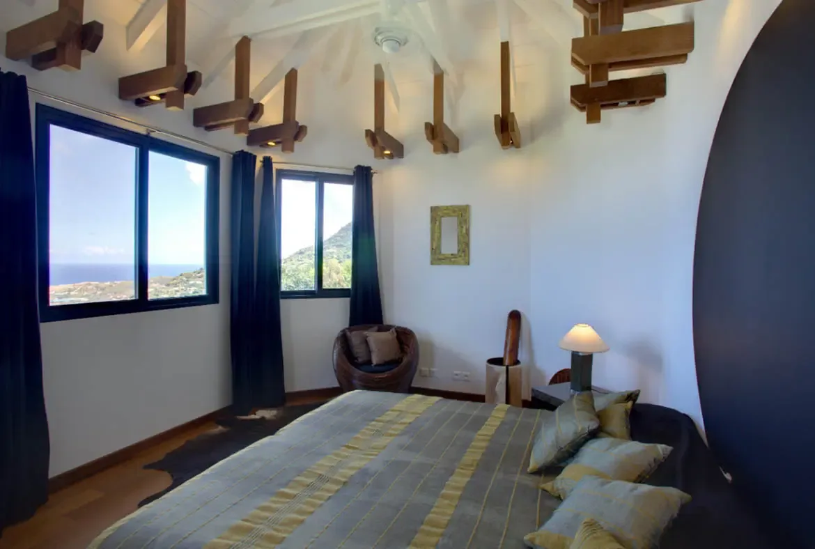 A curved dark accent wall and sage-green pillows frame a bed under sculptural ceiling brackets, where an ocean view and a papasan chair add coastal warmth.