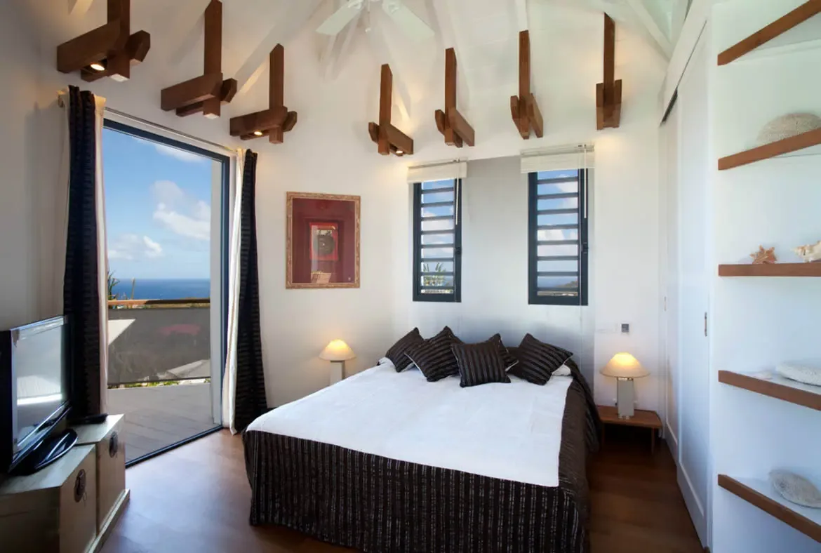 A white bed with brown pillows and a warm art piece sits under sculptural ceiling beams, where open glass doors lead to a deck overlooking turquoise ocean and a shelf displays seashells.
