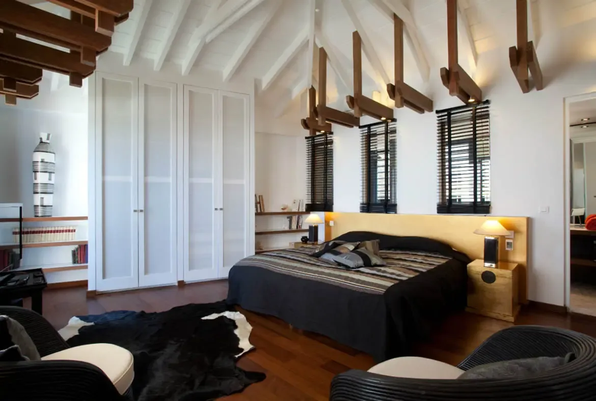 A wide wood headboard and charcoal bedding sit under hanging wooden ceiling beams, where wicker armchairs and a cowhide rug rest on dark hardwood beside a white wardrobe.