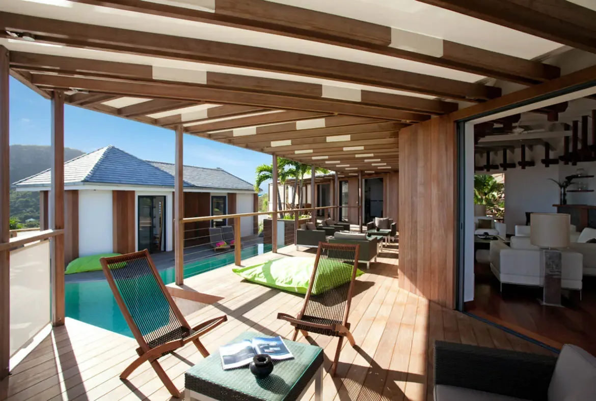Wooden loungers and a lime-green beanbag rest on a deck under a slatted pergola, where a turquoise pool stretches beside a white-walled villa toward a deep blue ocean.