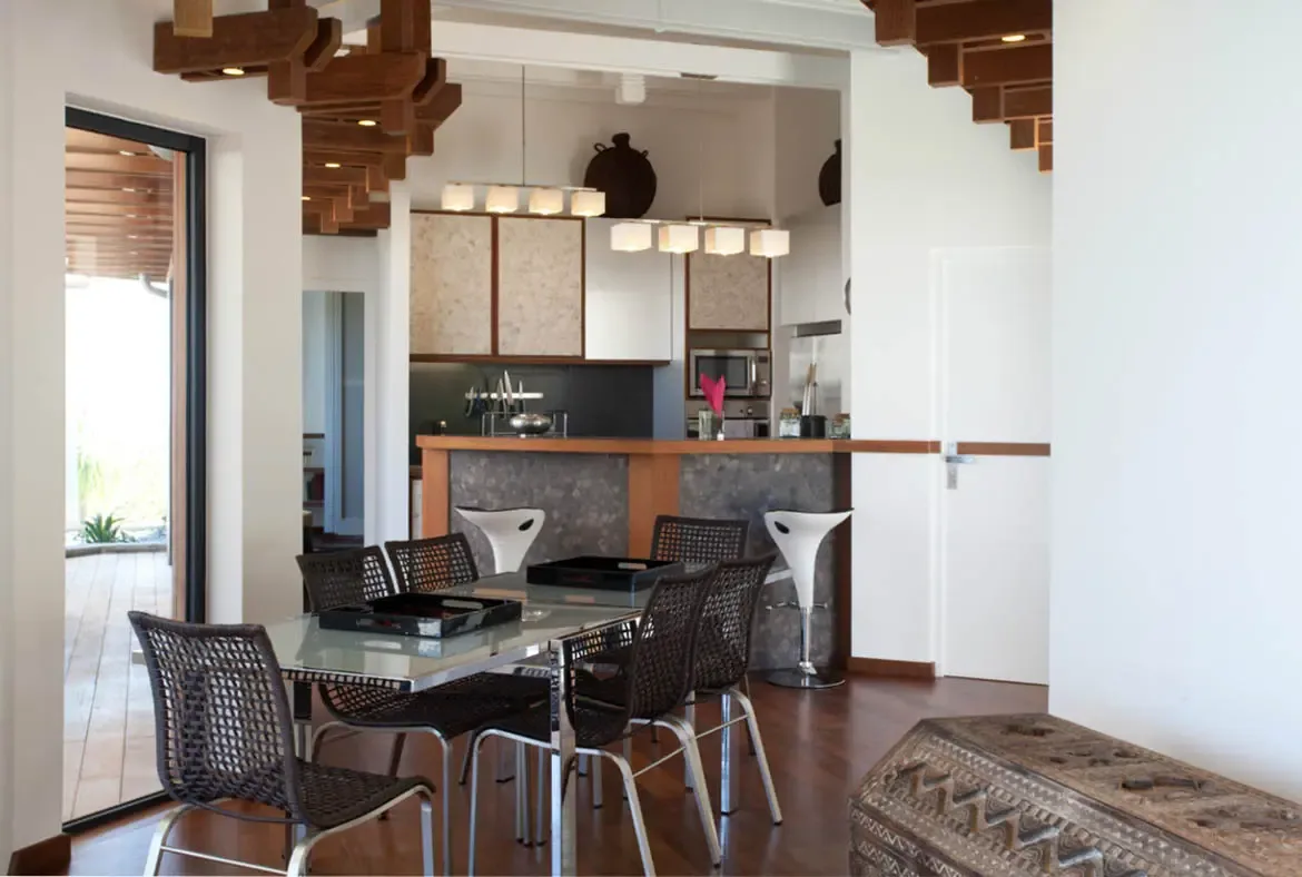 A glass dining table with woven chairs sits beside a river-stone breakfast bar and white stools, where sculptural ceiling beams and a carved wood chest add warmth to the open-concept space.