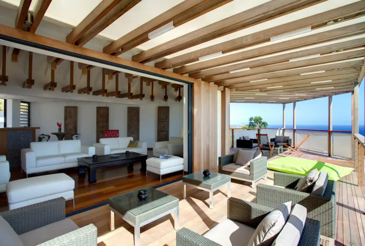 White leather sofas and carved wood panels anchor a living room under massive ceiling beams, where retracted glass doors connect to a deck with grey wicker seating and lime-green loungers overlooking the ocean.