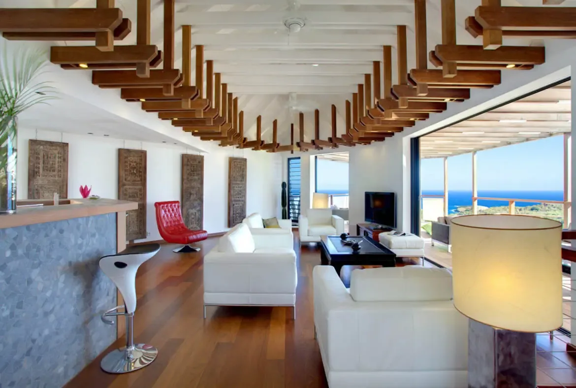 White leather sofas and a curved red armchair sit under cascading ceiling beams, where carved wood panels and a stone bar with white stools face open glass doors to the ocean.