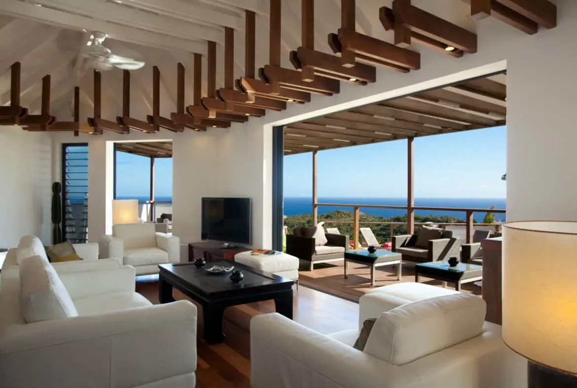 White leather sofas and a dark coffee table sit under sculptural ceiling beams, where retracted glass doors open to a shaded deck with an ocean panorama beyond cable railings.