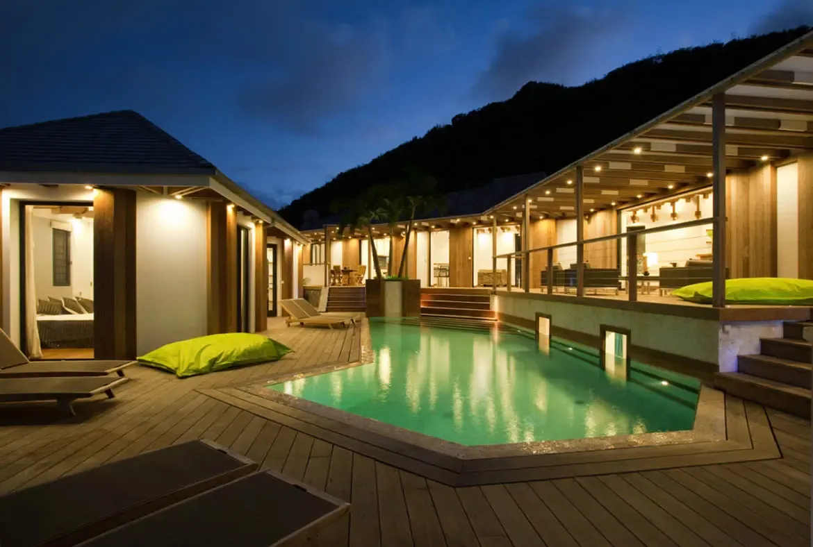 At twilight, a turquoise pool glows between warm-lit wooden pavilions, where lime-green beanbags and lounge chairs rest on a deck backed by a dark mountain silhouette.