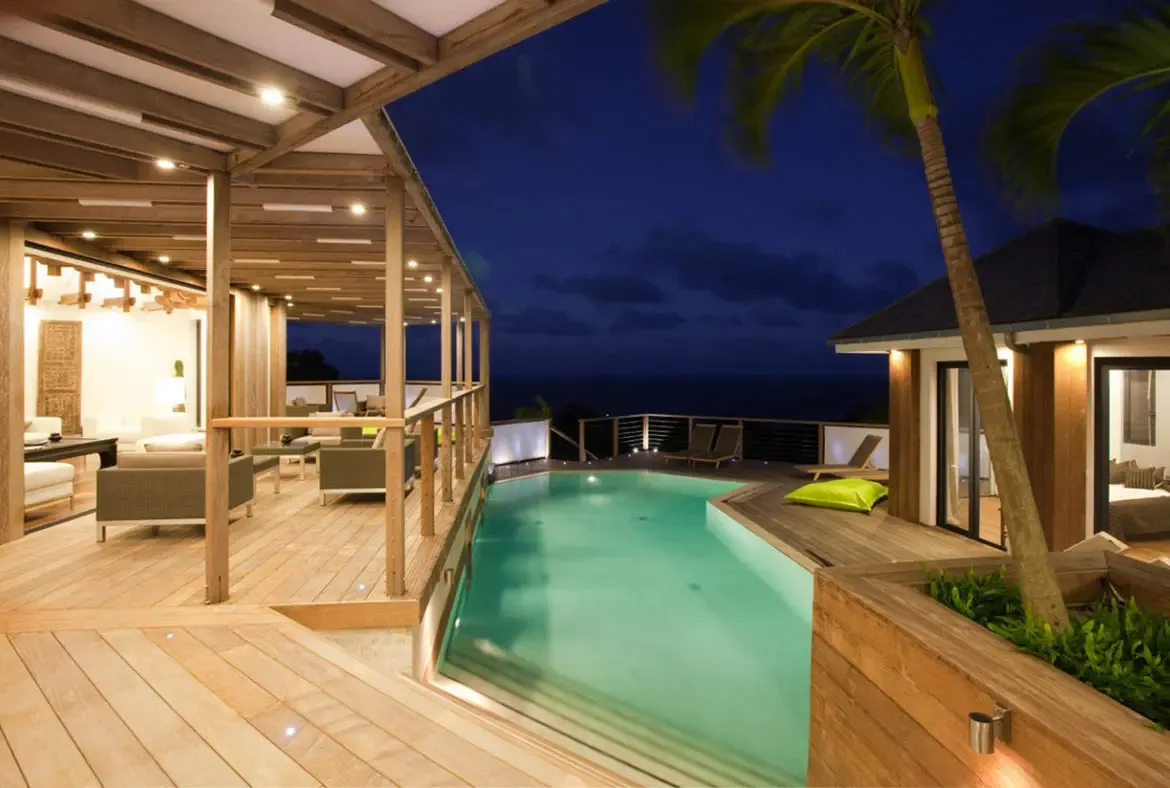 At dusk, a turquoise infinity pool glows beside a wooden pavilion, where a single lime-green lounger and palm trees frame an ocean view under an indigo sky.