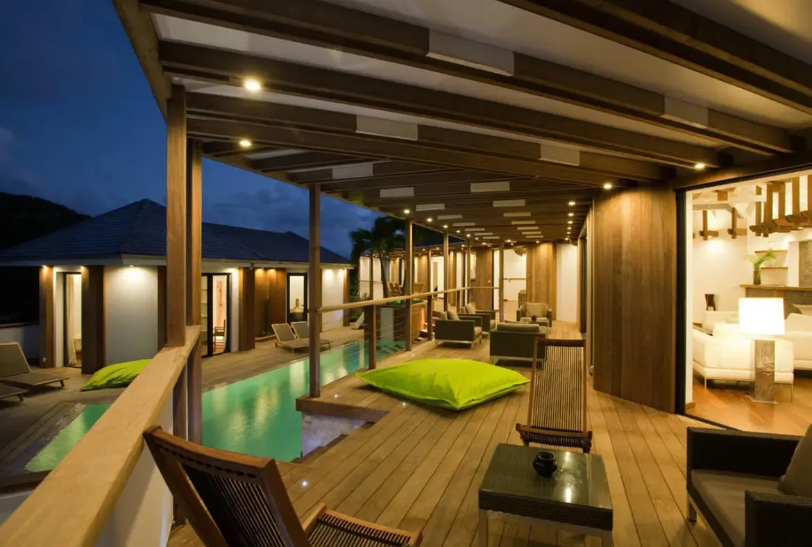 At blue hour, a glowing pool stretches along a covered wooden gallery, where recessed lights and warm interior glow illuminate multiple pavilion wings against an indigo sky.