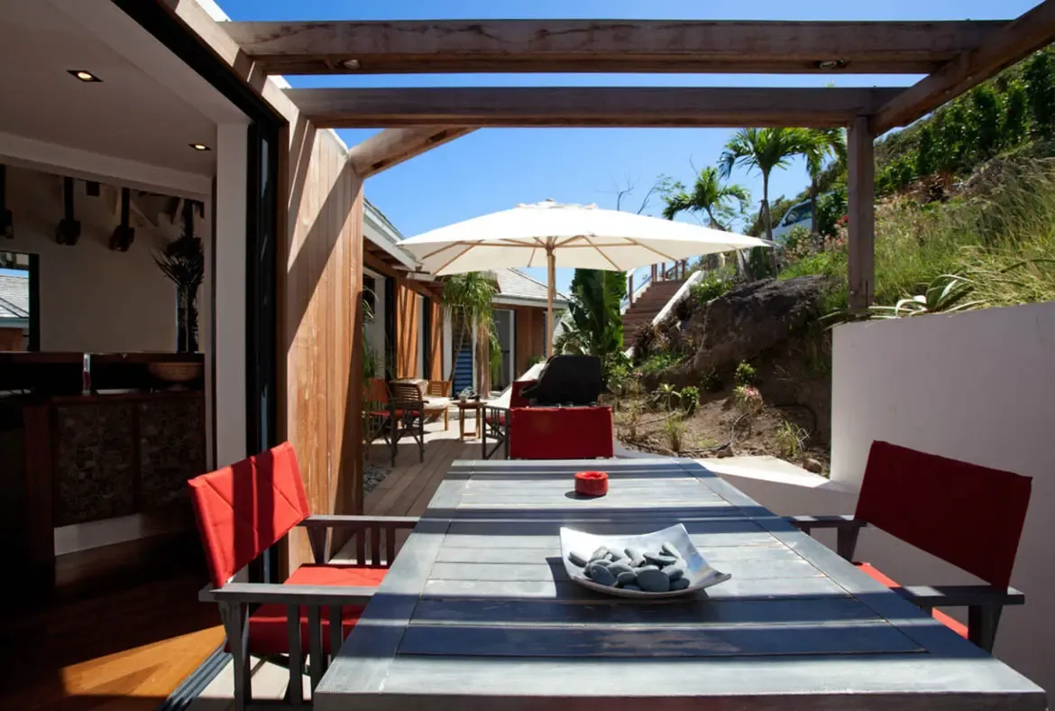 A weathered wood table with red chairs sits under a pergola casting geometric shadows, where glass doors open to a hillside deck with palm trees and a white umbrella.