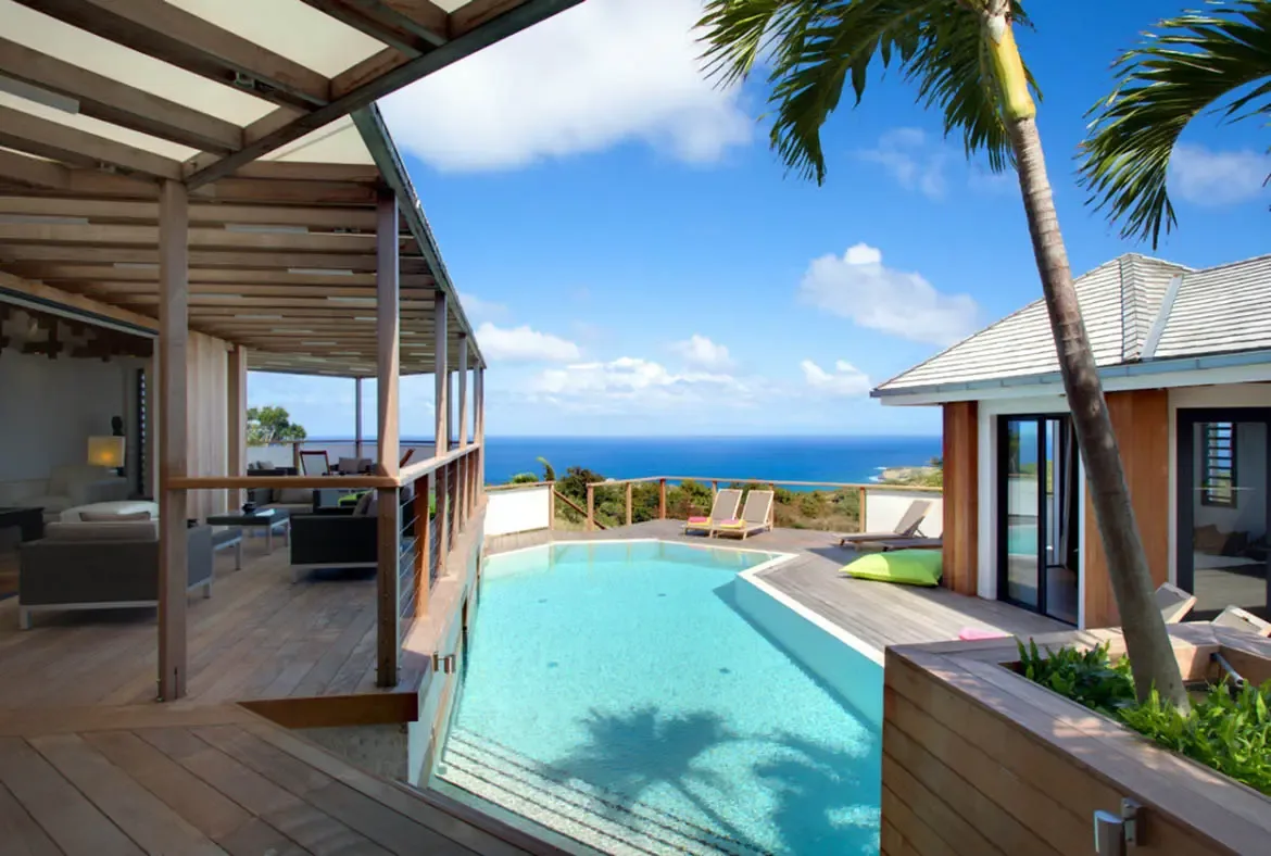 A geometric turquoise pool with shallow steps sits on a weathered deck, where lounge chairs with pink and yellow towels and a lime-green beanbag overlook a deep blue ocean panorama.