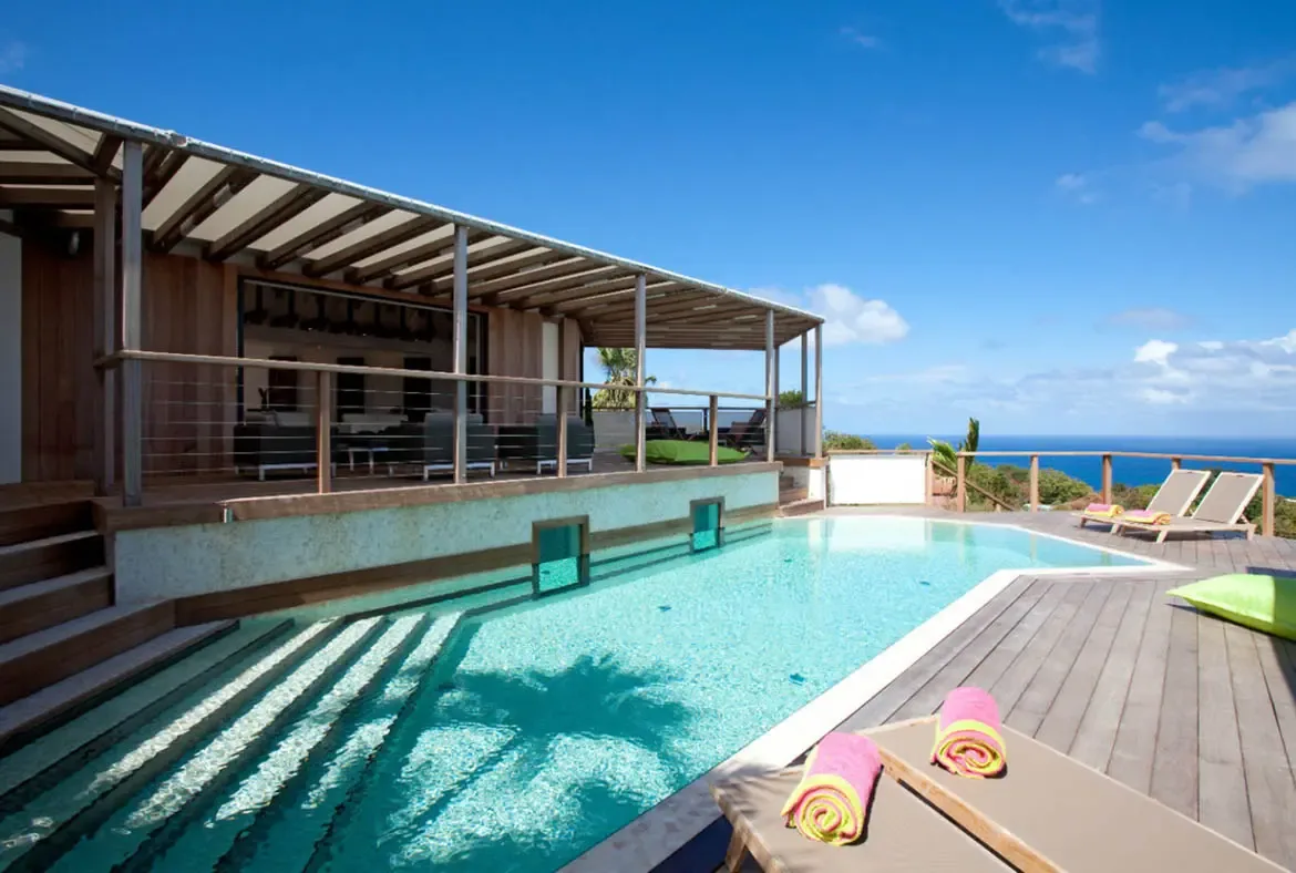 A light-blue pool with shallow steps sits on a gray deck under a slatted pergola, where rolled pink and yellow towels on loungers and a lime-green cushion face a sweeping ocean view.