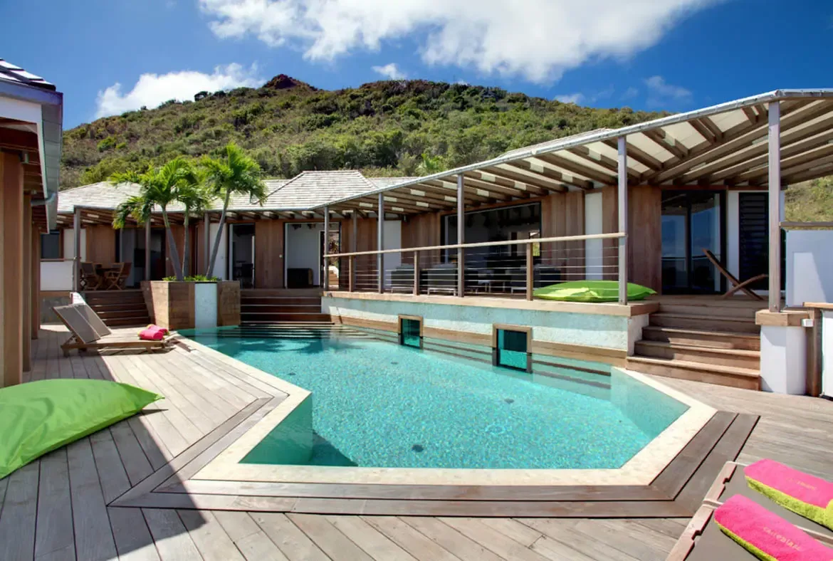 An angular turquoise pool with shallow steps sits on a geometric deck, where turquoise underwater windows and lime-green beanbags add playfulness beneath a slatted wood pergola and palm trees.