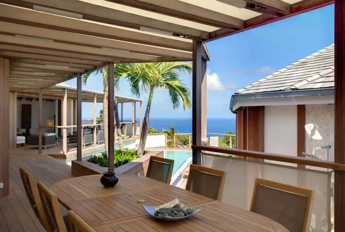 A slatted wood dining table and chairs sit under a pergola on a wide deck, where a pool edge and turquoise ocean stretch beyond palm trees under a clear sky.