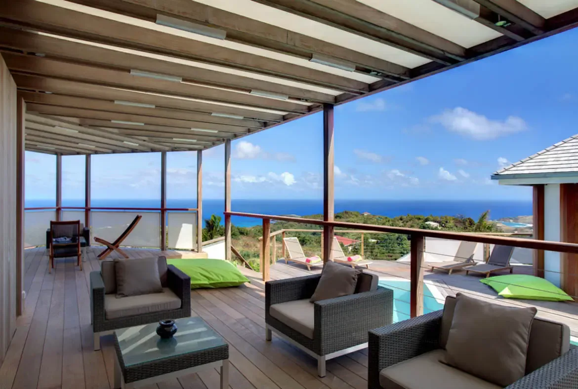 Grey wicker armchairs and lime-green floor cushions sit on a wooden deck under a slatted pergola, where cable railings frame a panoramic ocean view stretching to the horizon.