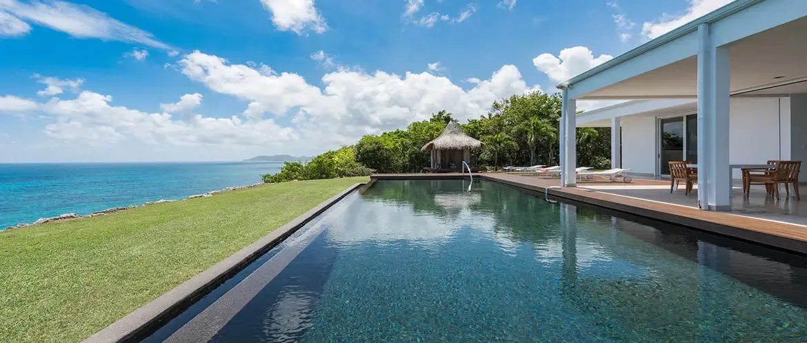 A rectangular infinity pool reflects the sky on a dark wood deck beside a white villa, where a green lawn, a thatched gazebo, and distant islands dot the turquoise-to-sapphire ocean.