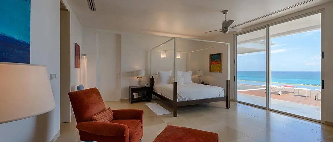 A white canopy bed with pendant lights faces floor-to-ceiling glass, where an infinity pool and turquoise ocean fill the view and a burnt-orange armchair adds warmth.