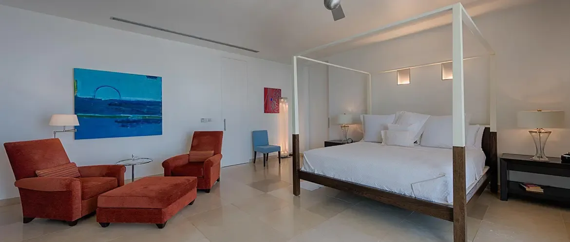 A white canopy bed with dark wood base sits on large-format tiles under a white ceiling, where burnt-orange armchairs and a deep blue abstract painting warm the minimalist room.