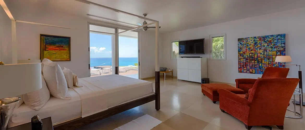 A dark wood canopy bed with white linens faces sliding glass doors, where burnt-orange armchairs and a colorful grid painting frame a panoramic turquoise ocean view.