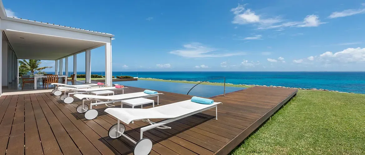 Four white wheeled loungers with coral and blue towels line a dark wood deck, where an infinity pool merges with a turquoise ocean above a manicured green lawn.