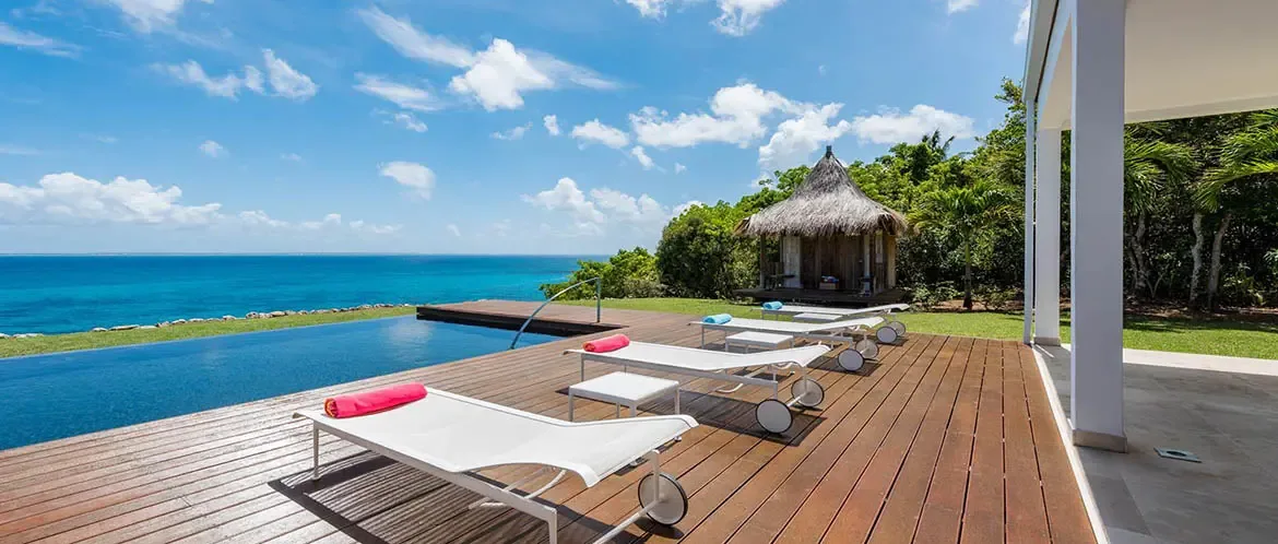 Four white loungers with pink and blue towels sit on a dark wood deck beside an infinity pool, where a thatched-roof gazebo nestles among palms above a turquoise ocean.
