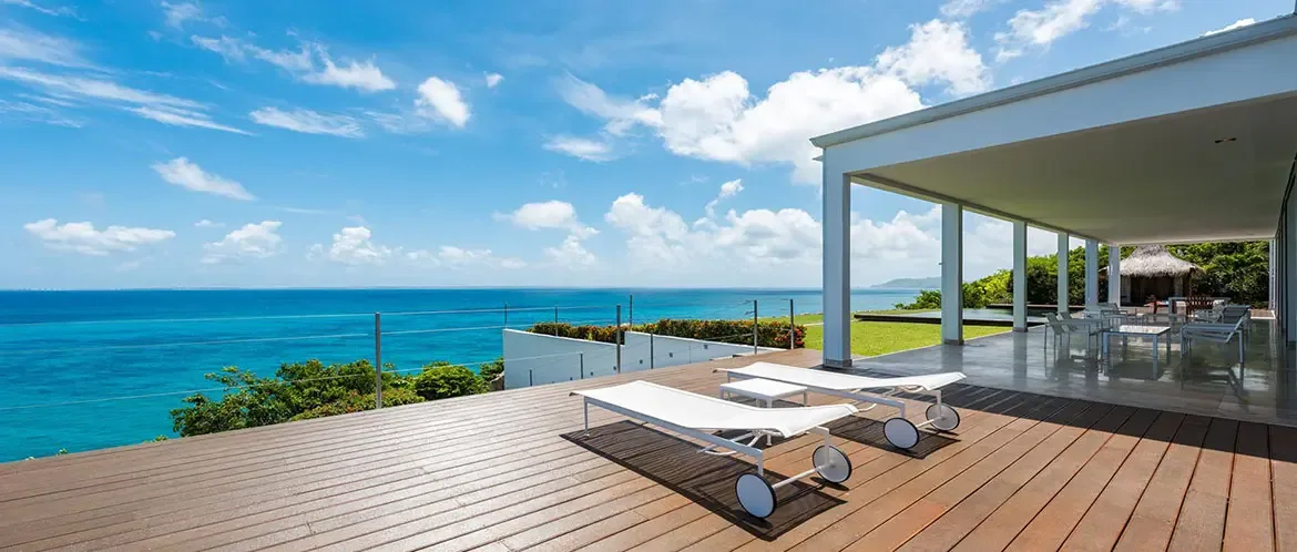 Two white wheeled loungers face a cable-railed deck above a hillside, where a white-pillared terrace and a thatched gazebo frame a turquoise-to-sapphire ocean panorama.