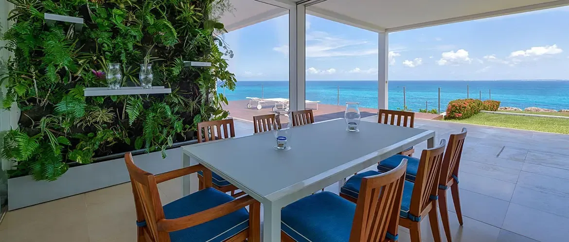 Blue upholstered chairs surround a grey dining table beside a lush vertical green wall, where glass panes frame loungers on a deck and a turquoise ocean stretches to the horizon.