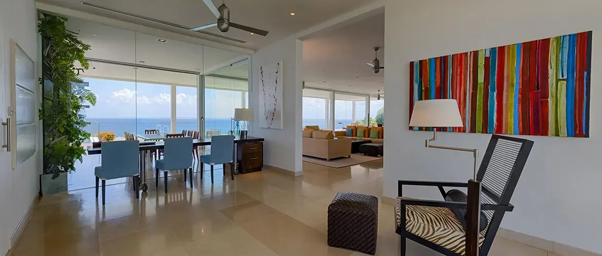Blue upholstered chairs surround a long dining table beside a lush vertical green wall, where a zebra chair and bold abstract art face floor-to-ceiling glass and a turquoise ocean panorama.