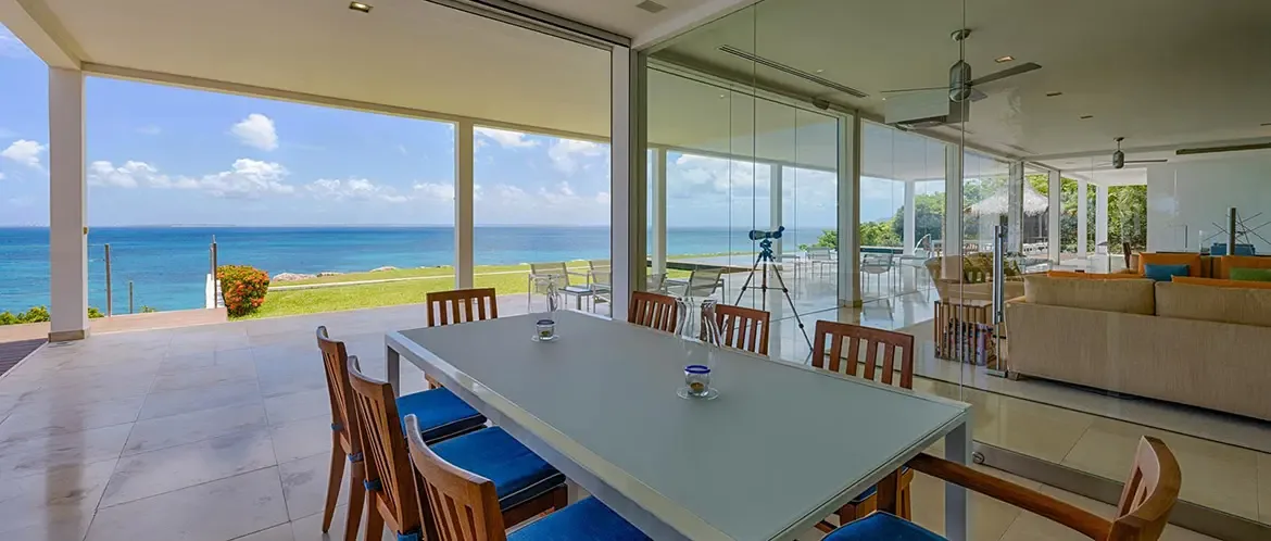 Blue chairs surround a white dining table on a covered terrace, where glass walls reveal a living room with tan sofas and a telescope aimed at the turquoise ocean.