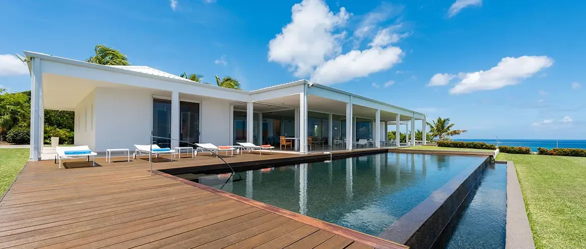 A white modern villa with glass doors sits beside a dark infinity pool on a wooden deck, where a manicured lawn and tropical palms overlook a turquoise ocean.