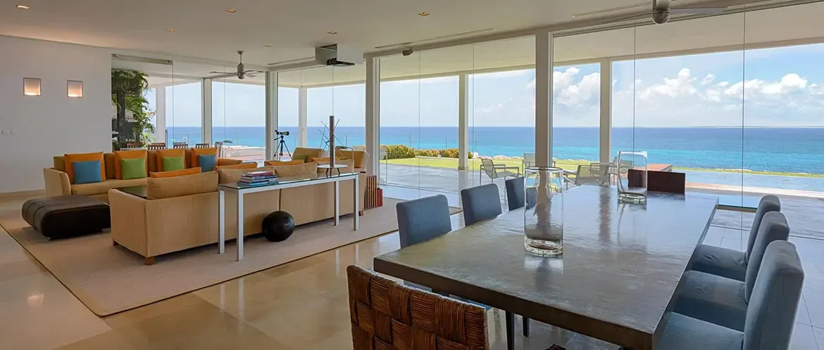 Tan sectional sofas with citrus pillows and a leather ottoman sit on a light rug, where a stone dining table with grey chairs and glass vases faces floor-to-ceiling glass and a turquoise ocean.