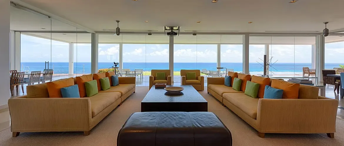 Two tan sofas with orange, lime, and turquoise pillows face a glass coffee table under a white ceiling with a projector, where floor-to-ceiling glass frames a panoramic turquoise ocean.
