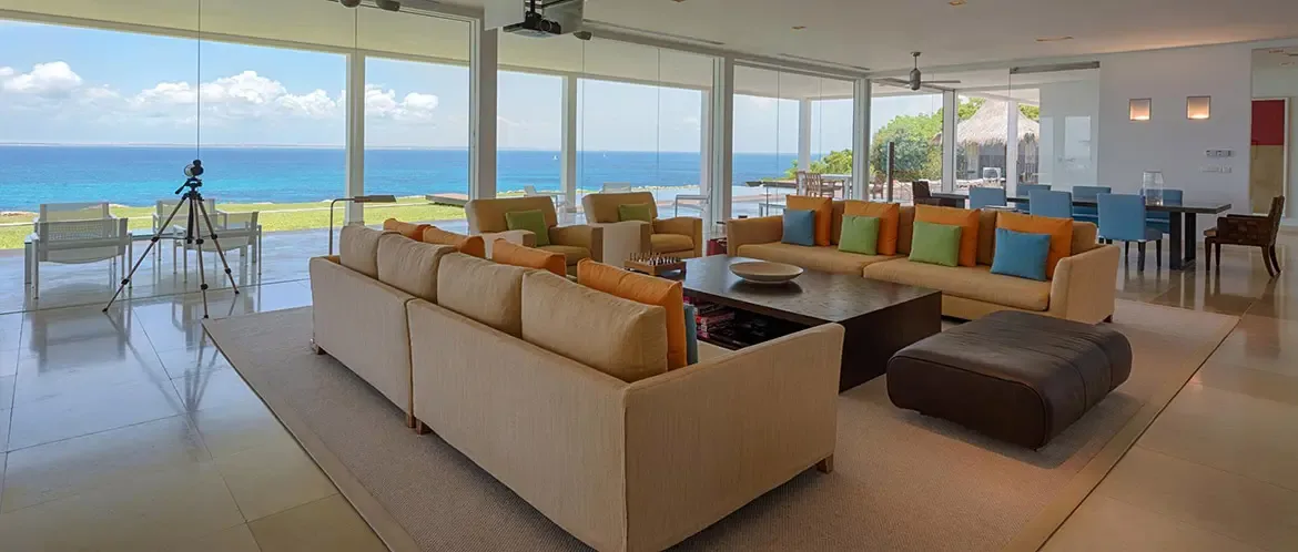 Two tan sectionals with turquoise and orange pillows face each other across a dark wood coffee table, where a telescope by the glass doors surveys the ocean and a terrace with white chairs.