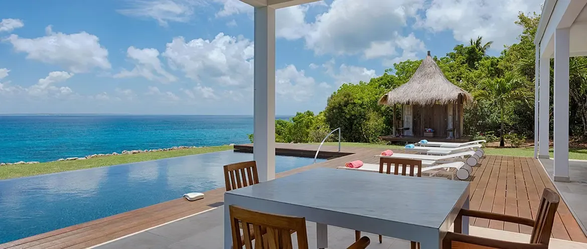 A grey dining table with wooden chairs sits under white pillars on a dark wood deck, where an infinity pool and a thatched gazebo overlook a turquoise ocean under scattered clouds.