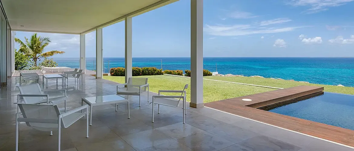 Minimalist white armchairs sit on polished stone tiles under white pillars, where a manicured lawn with red flowers leads past an infinity pool to a turquoise ocean horizon.