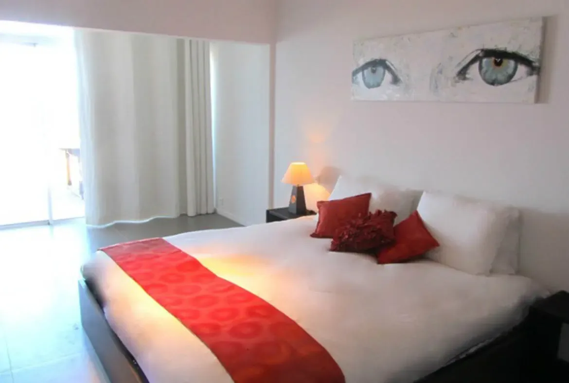 Surrender to tranquility in Agave Azul's elegantly styled bedroom, where bold red accents and a captivating eye-themed artwork blend with soft natural light streaming through sheer curtains. Step onto your private patio just beyond the glass doors and let St. Barths' serene energy envelop your stay.                                                                                                                                                                                            +
 