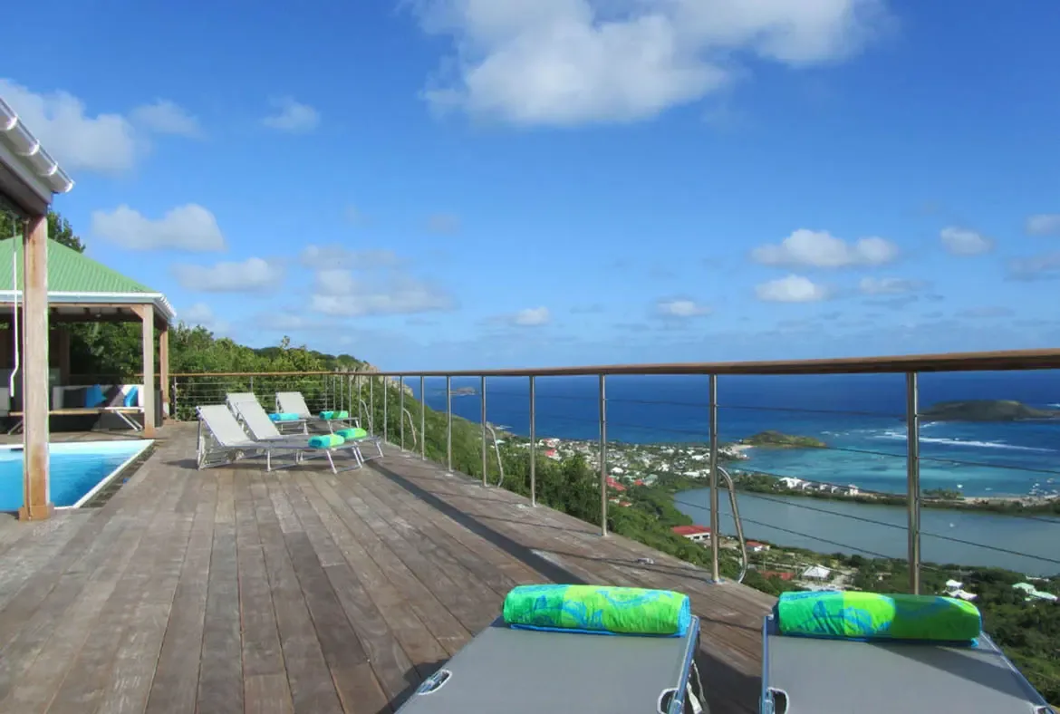 Soak in the Caribbean sunshine on Villa Agave Azul's expansive wooden deck, where plush lounge chairs and vibrant turquoise towels beckon you to unwind. Sip a tropical drink while gazing at the endless blue sea, with the island's lush coastline and distant islets unfolding beneath a cloud-dappled sky.                                                                                                                                                                                                         +
 