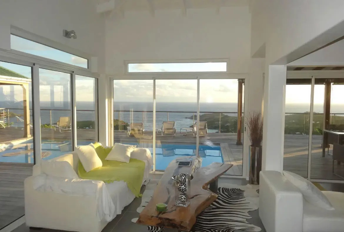 Bask in the golden hour glow at Villa Agave Azul, where contemporary design meets the boundless beauty of St. Barths. This sun-drenched living area seamlessly blends indoor luxury with the sparkling azure of your private pool and the panoramic Caribbean horizon beyond. Every detail, from the bespoke live-edge wood table to the vibrant citrus accents, invites you to unwind in true island sophistication.                                                                                               +
 