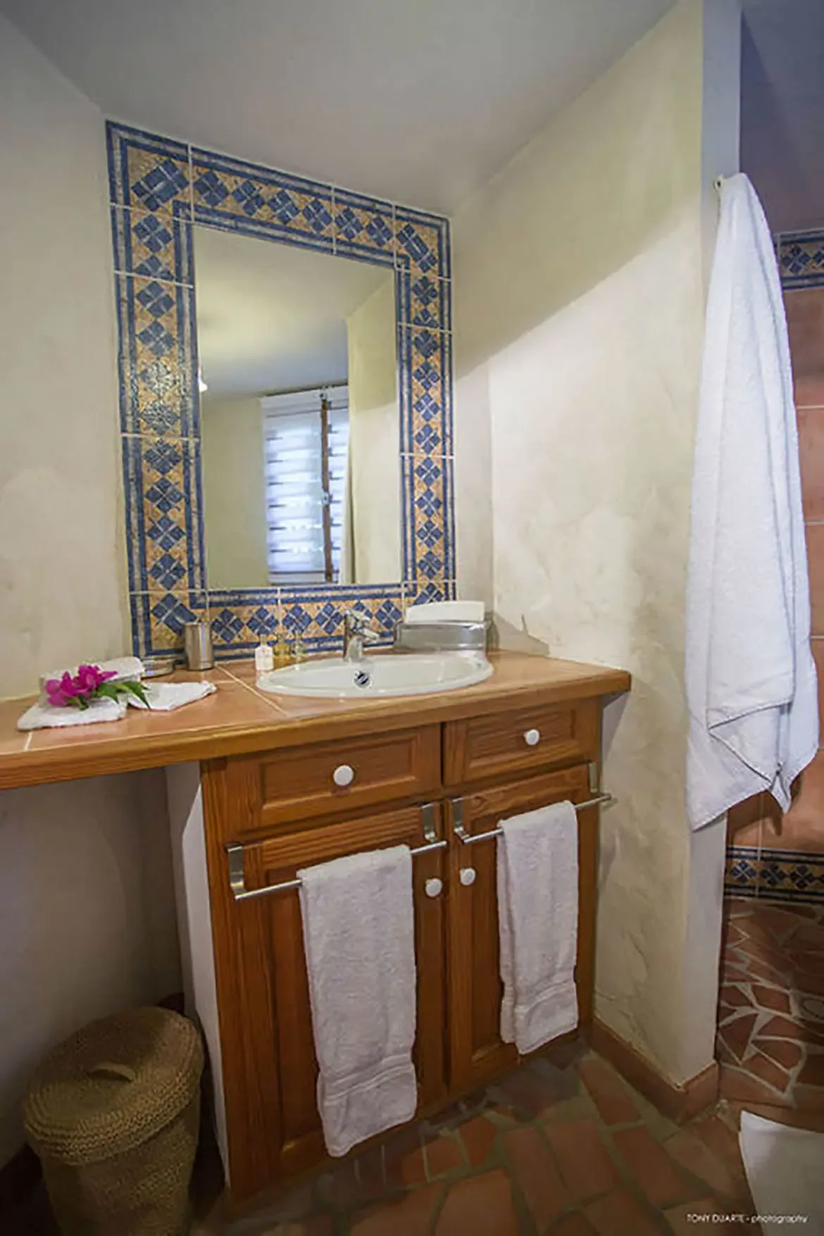 Step into this St. Barts villa bathroom where hand-painted blue and gold tilework frames the mirror above a warm wooden vanity, blending Caribbean charm with refined comfort. Fresh orchids and plush white towels invite you to savor every moment of your tropical retreat.