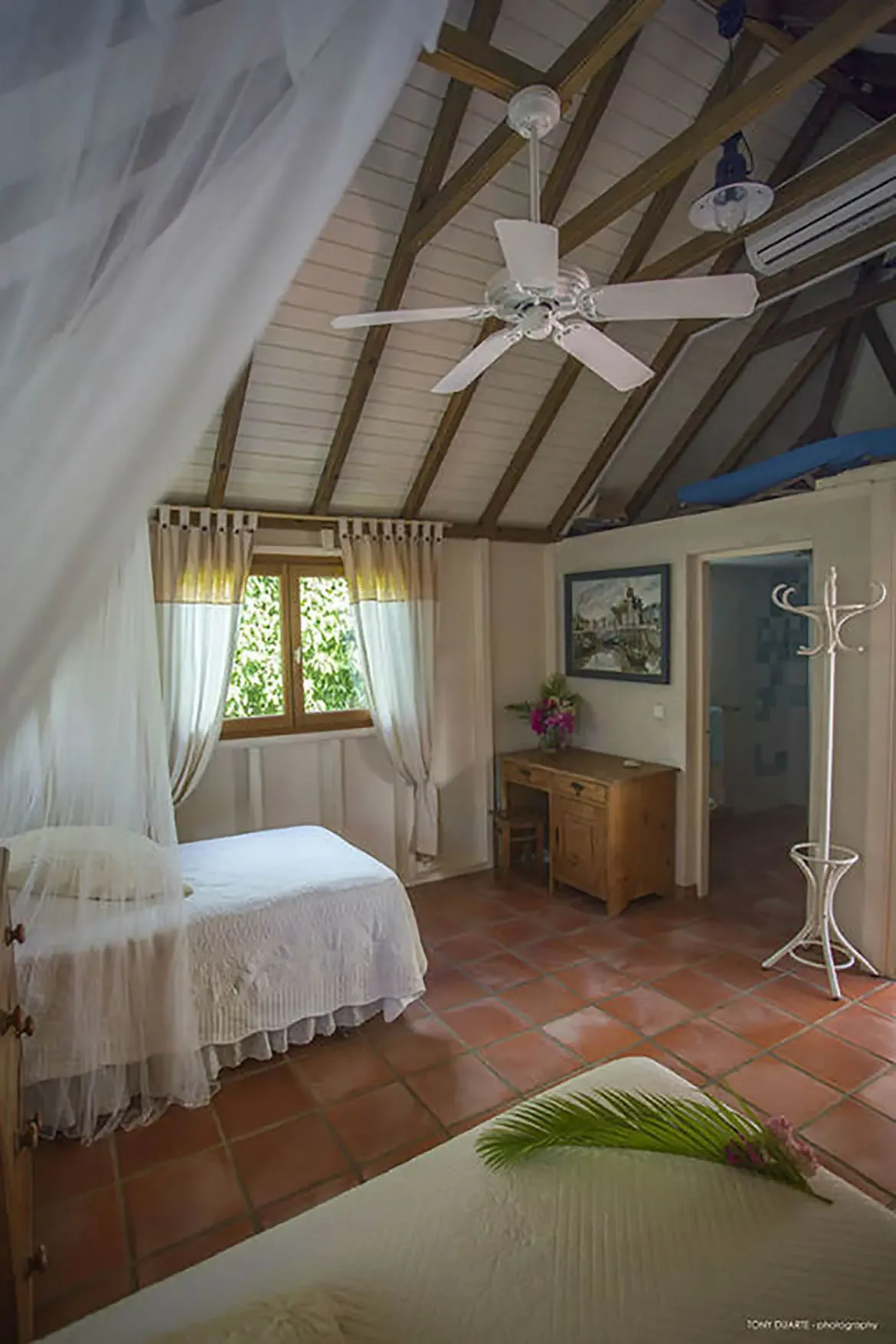 Drift into relaxation in this airy St. Barths retreat, where a billowing canopy bed meets natural light filtering through lush green windows. Rustic wooden beams and warm terracotta floors blend with modern comforts to create a serene escape for restorative rest.