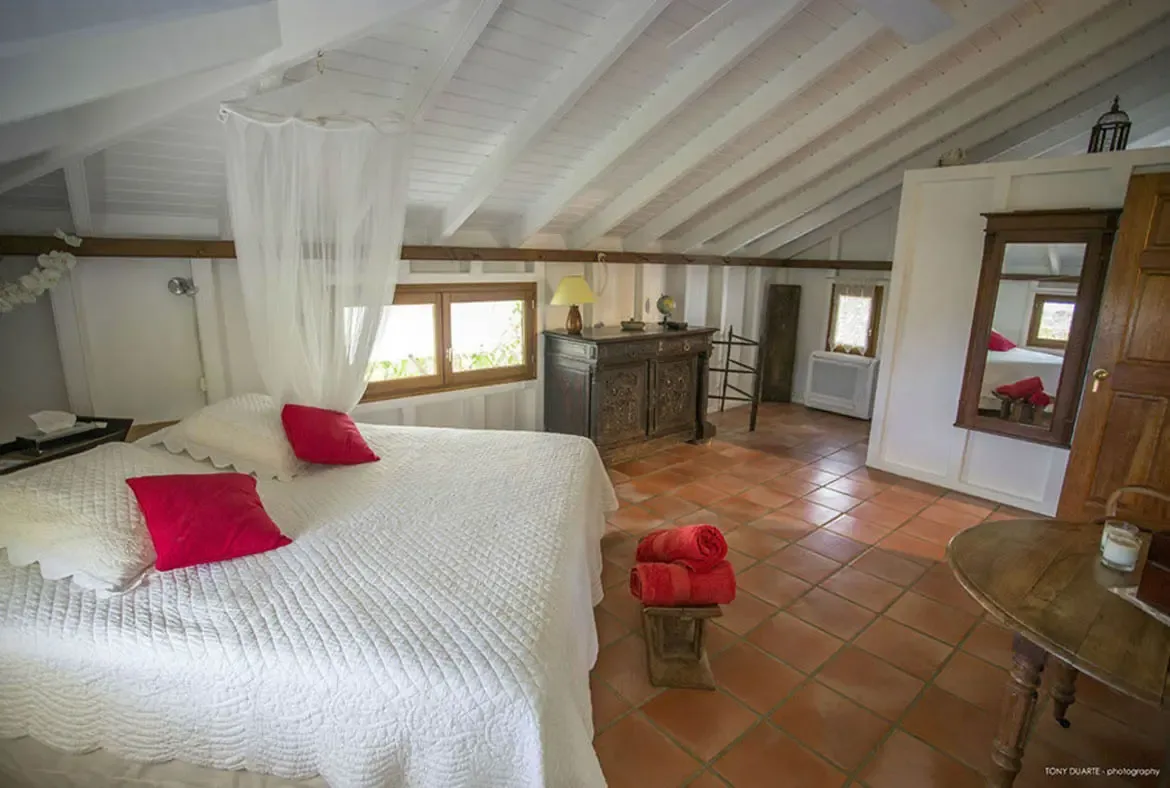 Unwind in this sunlit St. Barths retreat, where a flowing white canopy drapes over a plush bed adorned with vibrant red accents, and terracotta floors blend seamlessly with exposed wooden beams for rustic elegance. Wake to natural light filtering through the window and the tranquil promise of island living in this thoughtfully designed sanctuary.