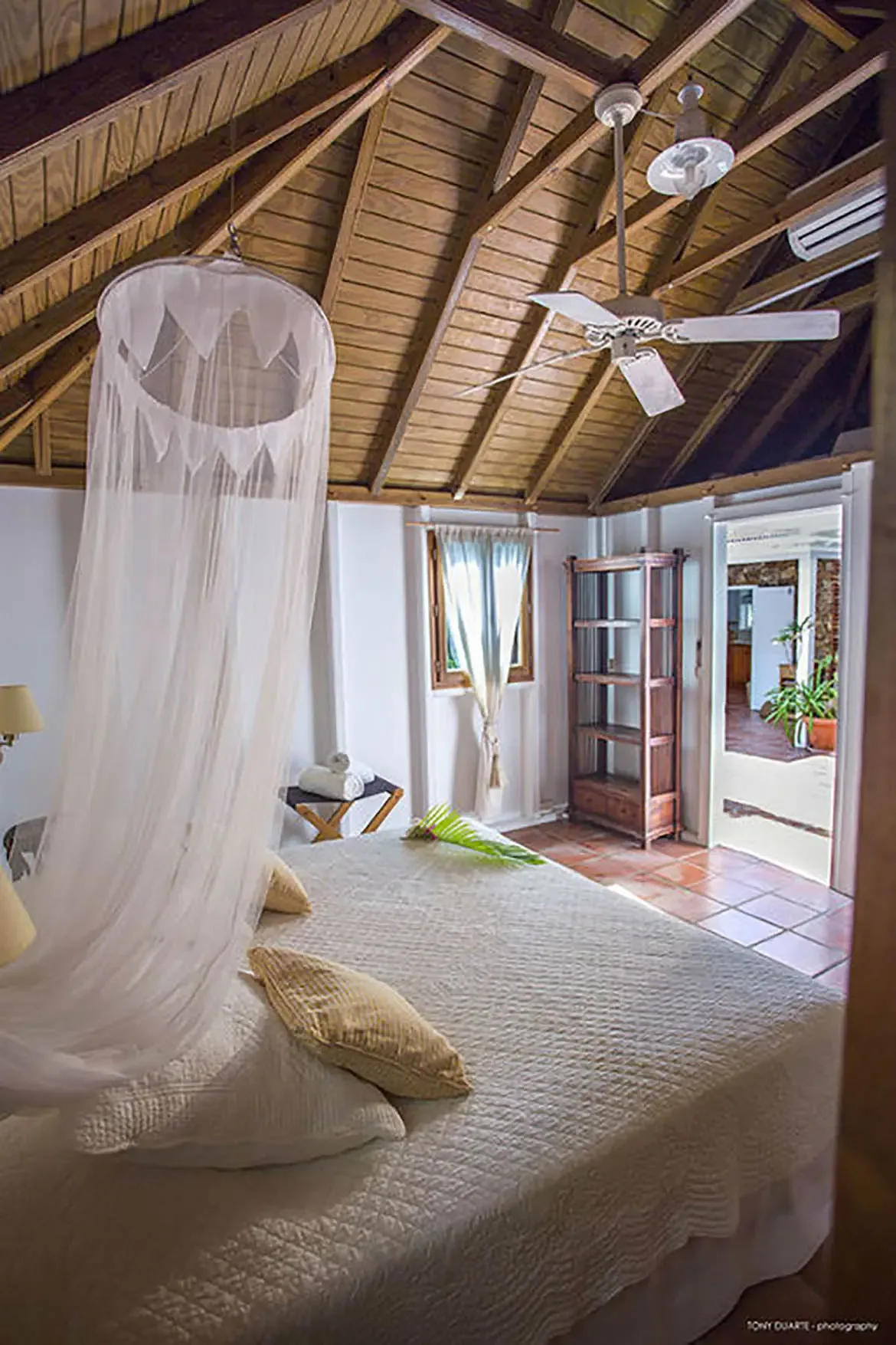 Drift into restful slumber beneath a flowing mosquito net canopy in this sunlit St. Barths bedroom, where rustic wooden beams and soft natural light blend with effortless island elegance. Step through the open doorway to a sun-drenched patio, inviting you to savor the seamless indoor-outdoor luxury of villa life.
