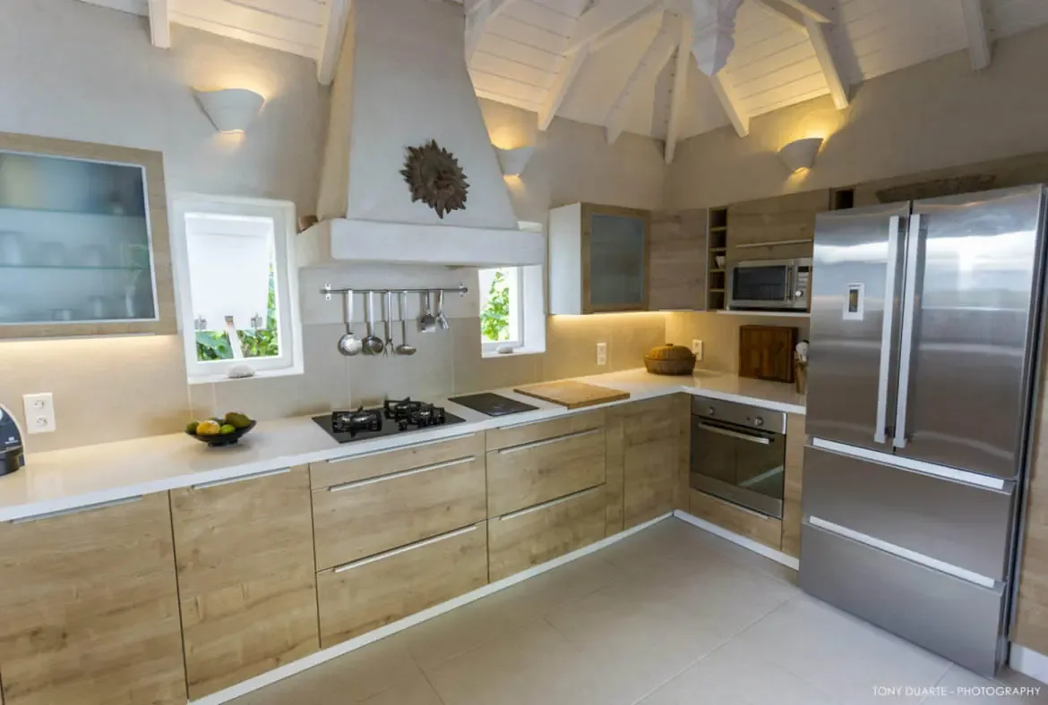 Craft culinary masterpieces in this sunlit kitchen, where sleek stainless steel appliances meet warm wood cabinetry under a vaulted ceiling. With natural light spilling through windows framing lush greenery, every meal feels like a seamless blend of luxury and island tranquility.