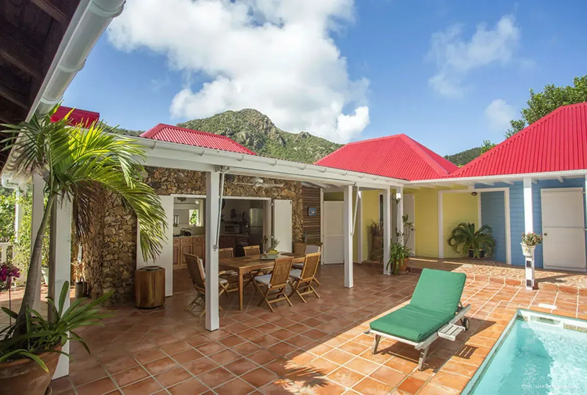 Savor the serenity of your private patio, where a sunlit lounge chair invites relaxation beside a glistening pool, with panoramic mountain vistas and vibrant red-roofed villas as your backdrop. Dine al fresco at the shaded wooden table, surrounded by tropical greenery and the warm St. Barths breeze.