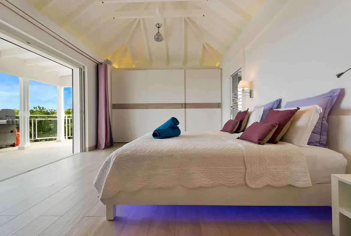 Unwind in this sun-drenched St. Barths bedroom, where a plush quilted bed invites rest beneath a vaulted ceiling. Step onto the private terrace to embrace views of lush greenery and the bright blue sky, then return to the soft glow of ambient lighting for a peaceful night's sleep.                                                                                                                                                                                                                     +
 