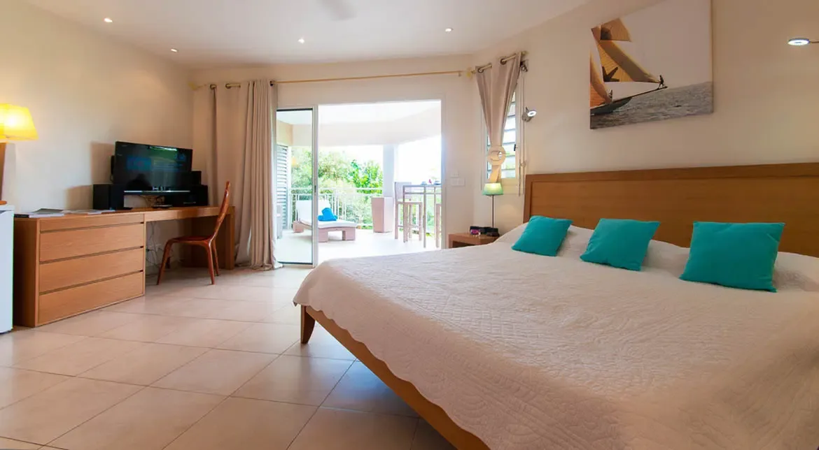 Step into this serene St. Barths bedroom, where a king-sized bed with crisp linens and vibrant turquoise accents awaits, while sliding glass doors reveal a private terrace framed by tropical greenery. Modern comforts like a sleek workspace and flat-screen TV blend seamlessly with the villa's airy, sunlit design for effortless island living.                                                                                                                                                       +
 