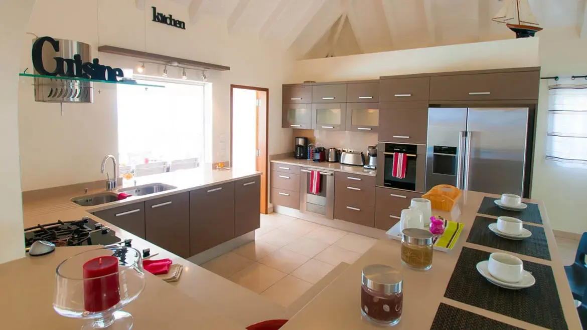 Savor sunrise coffee at the spacious kitchen island in this St. Barths villa, where sleek cabinetry and stainless steel appliances blend with sunlit vaulted ceilings. Prepare gourmet meals with ease, then enjoy them at the dining counter adorned with fresh tableware and vibrant accents.                                                                                                                                                                                                         +
 