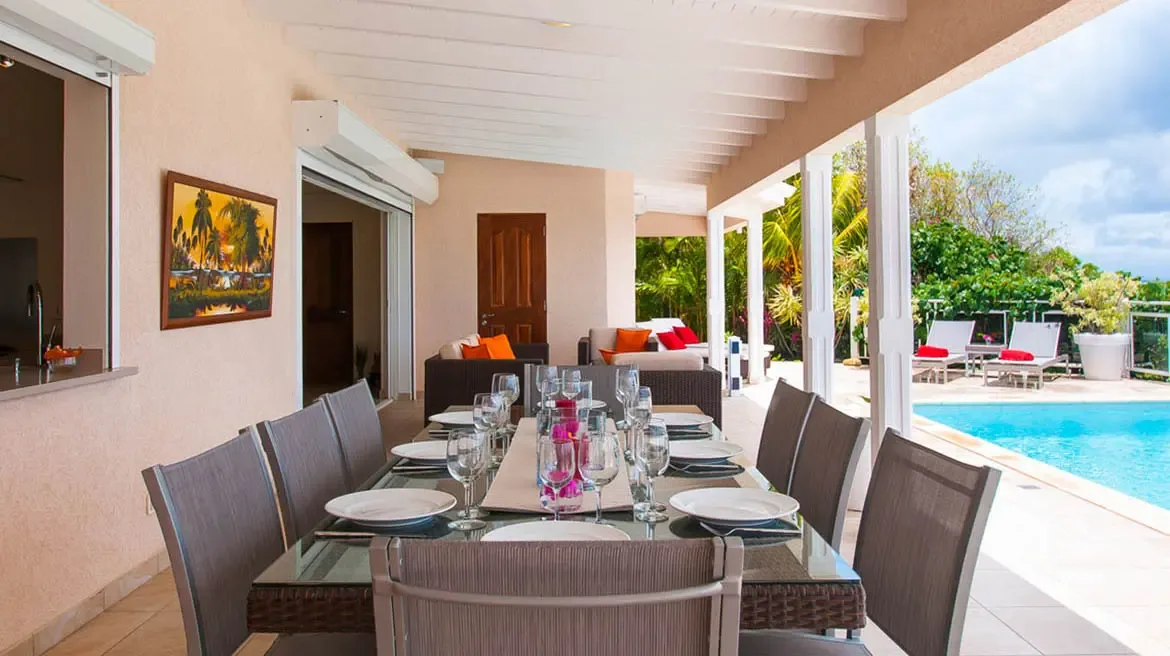 Gather for elegant al fresco meals beneath the covered terrace of Villa Au Coeur du Rocher, where a polished glass dining table and woven chairs frame panoramic views of the turquoise pool and tropical gardens. The seamless blend of indoor-outdoor living, with plush lounge seating and vibrant island art, creates an effortless luxury for sharing meals with loved ones.                                                                                                                              +
 