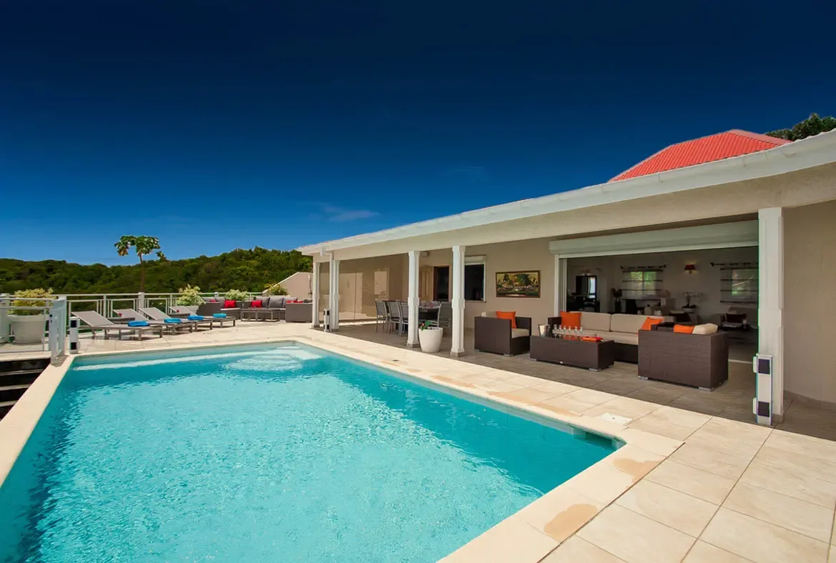Discover serene luxury at Villa Au Coeur du Rocher, where a glistening pool meets a spacious covered living area framed by St. Barths' lush hills and endless blue sky. Unwind on sun-drenched lounge chairs or gather in the open-air lounge, designed for effortless island living with seamless indoor-outdoor flow.                                                                                                                                                                                  +
 