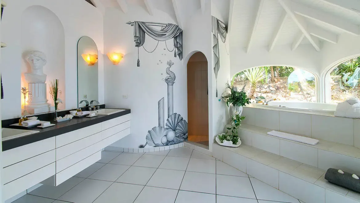Unwind in the artfully designed bathroom of Villa Byzance, where a hand-painted mural and sunken tub blend luxury with coastal inspiration. The double vanity, arched windows framing lush island greenery, and bespoke details create a serene sanctuary for relaxation.                                                                                                                                                                                                                             +
 