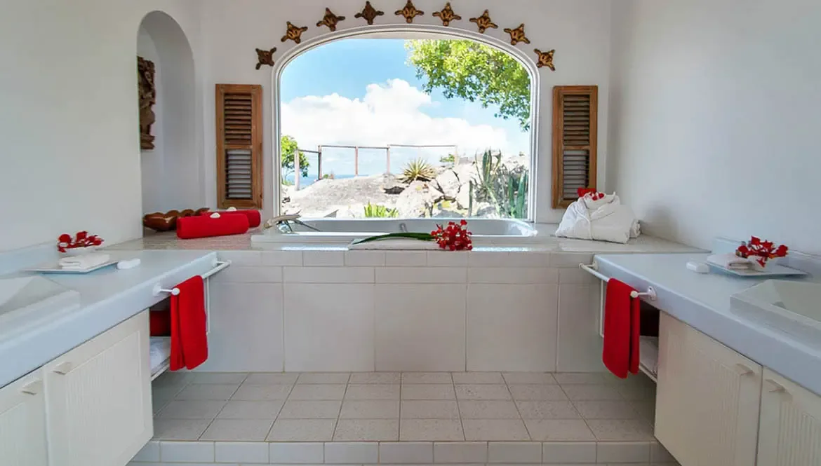 Unwind in Villa Byzance's sunlit bathroom, where a freestanding tub frames sweeping ocean vistas and tropical greenery. Vibrant red accents and artisanal star-shaped tilework elevate the space into a serene sanctuary, merging luxury with St. Barths' natural beauty.                                                                                                                                                                                                                                               +
 