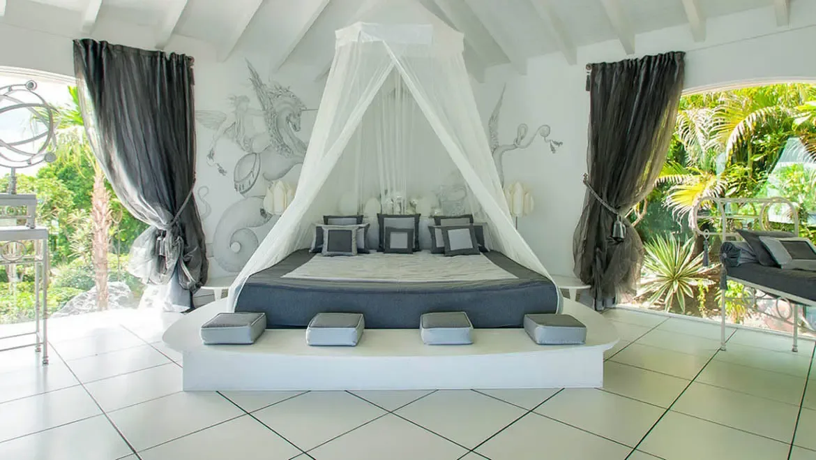 Drift into serenity beneath a billowing white canopy bed, where hand-painted griffin murals and soft gray accents create a dreamy escape. Floor-to-ceiling windows dissolve boundaries between the airy interior and lush tropical gardens, inviting the rhythm of St. Barts into your private sanctuary.                                                                                                                                                                                                      +
 